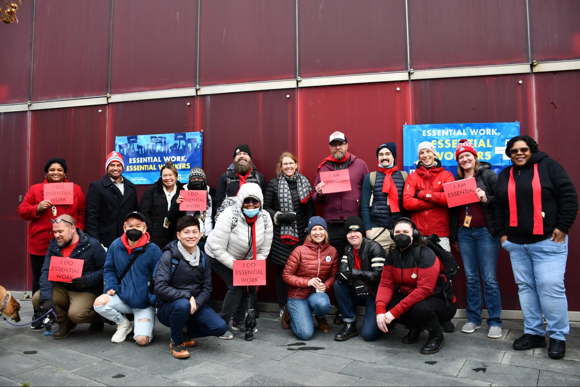 A group of people posing together outside in front of a red wall holding red signs for an essential workers demonstration.