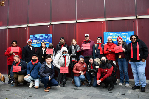 A group of people posing together outside in front of a red wall holding red signs for an essential workers demonstration.