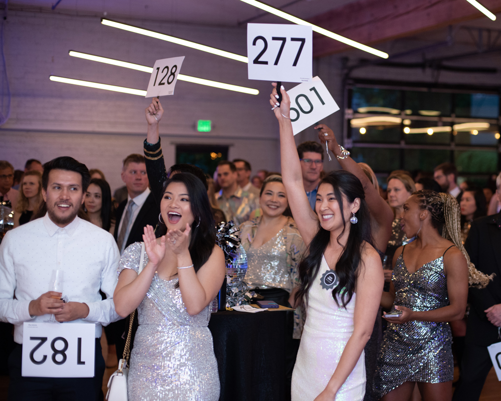 Young professionals cheer and raise numbered paddles at a formal fundraising gala