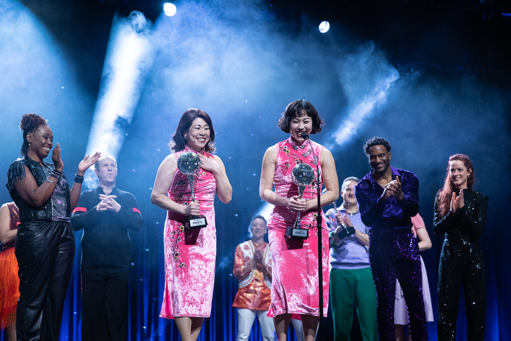 Dancer pairs gathered on stage cheer as two dancers accept mirrorball trophies.