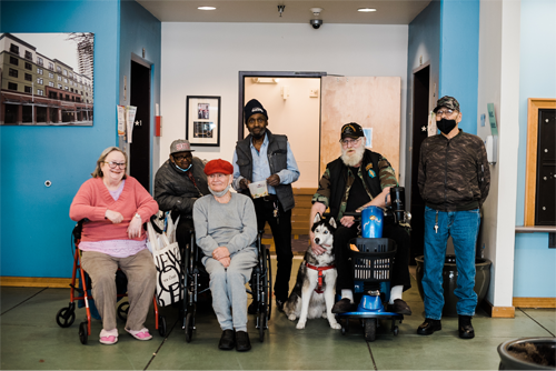 Five individuals are positioned across the room, two of whom are seated in wheelchairs, while another stands with a service dog. To the side, one person operates a scooter. The room has a vibrant blue wall and is adorned with framed pictures, while a large photograph of building exteriors hangs on an adjacent wall.