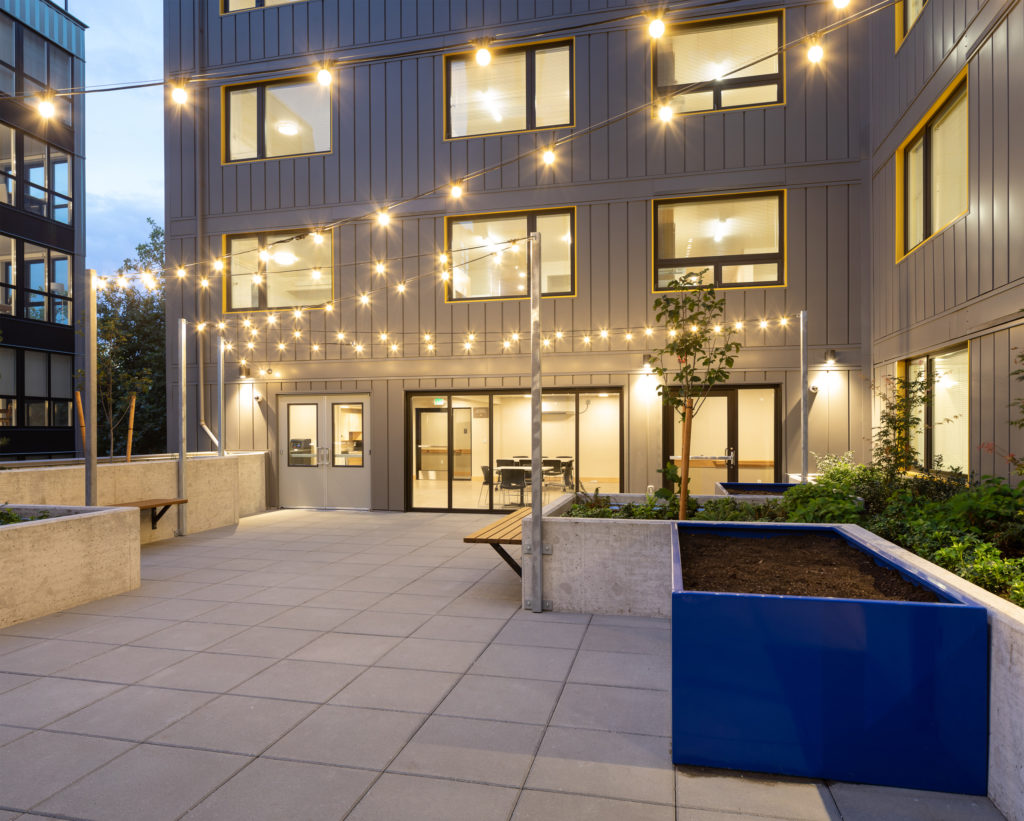 Modern courtyard with string lights, surrounded by buildings with large windows, at dusk.