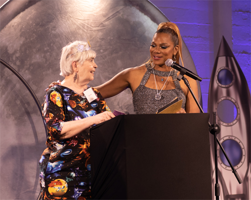 A woman proudly holds an award while standing beside an older woman, symbolizing recognition and respect.