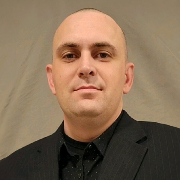A bald man in a black shirt and jacket, looking directly at the camera with a neutral expression.