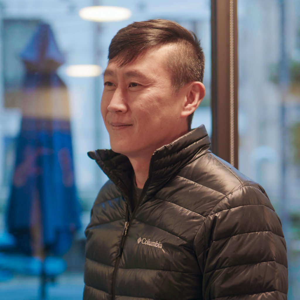 A man in a black Columbia jacket smiling slightly while standing indoors near a window