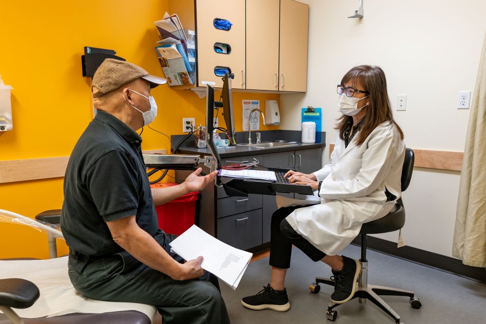 A healthcare provider speaking with a patient in a medical office. Both are wearing face masks.