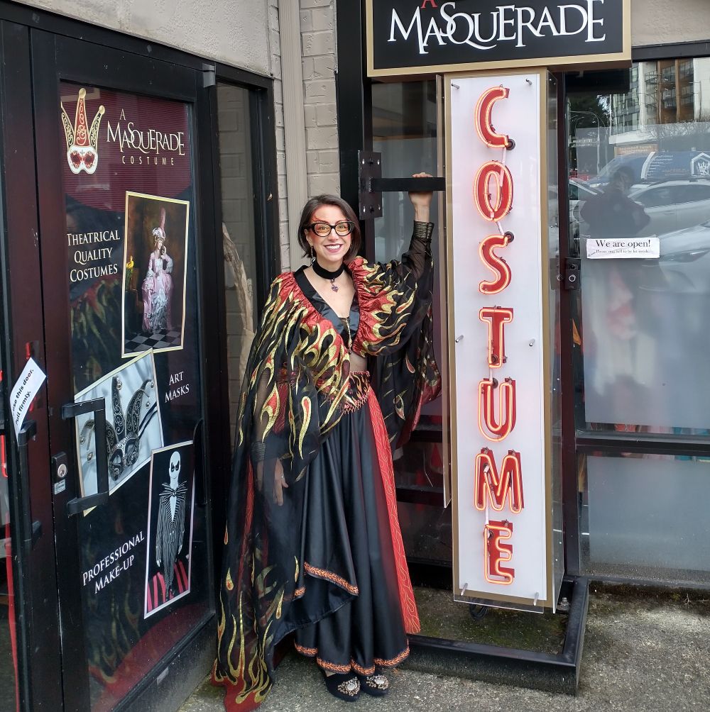 A person in a red and black dress with red and yellow flames across the top of the dress is standing in front of a costume shop