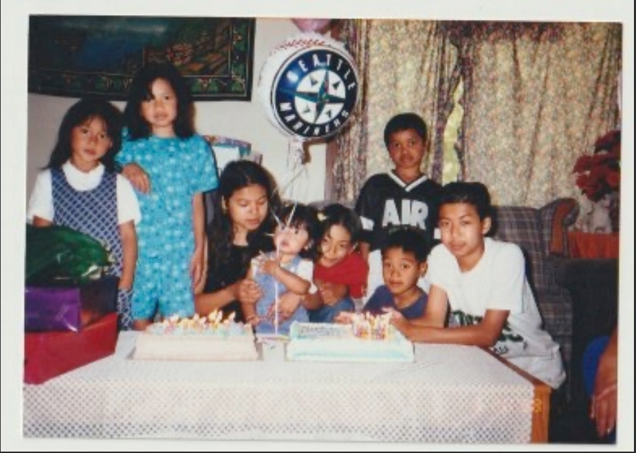 A group of young family members around a table with two cakes, with a Seattle Mariners balloon in the middle